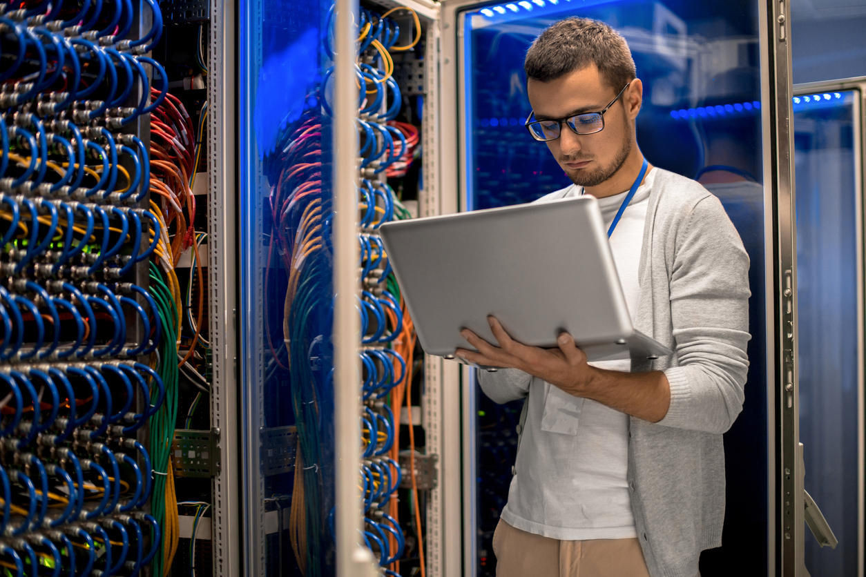 A computer support specialist consults a laptop while standing in a server room.