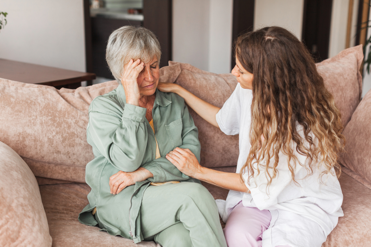 Young Woman Sitting on a Couch With an Elderly Woman Who Is Holding Her Head Trying to Remember Something.