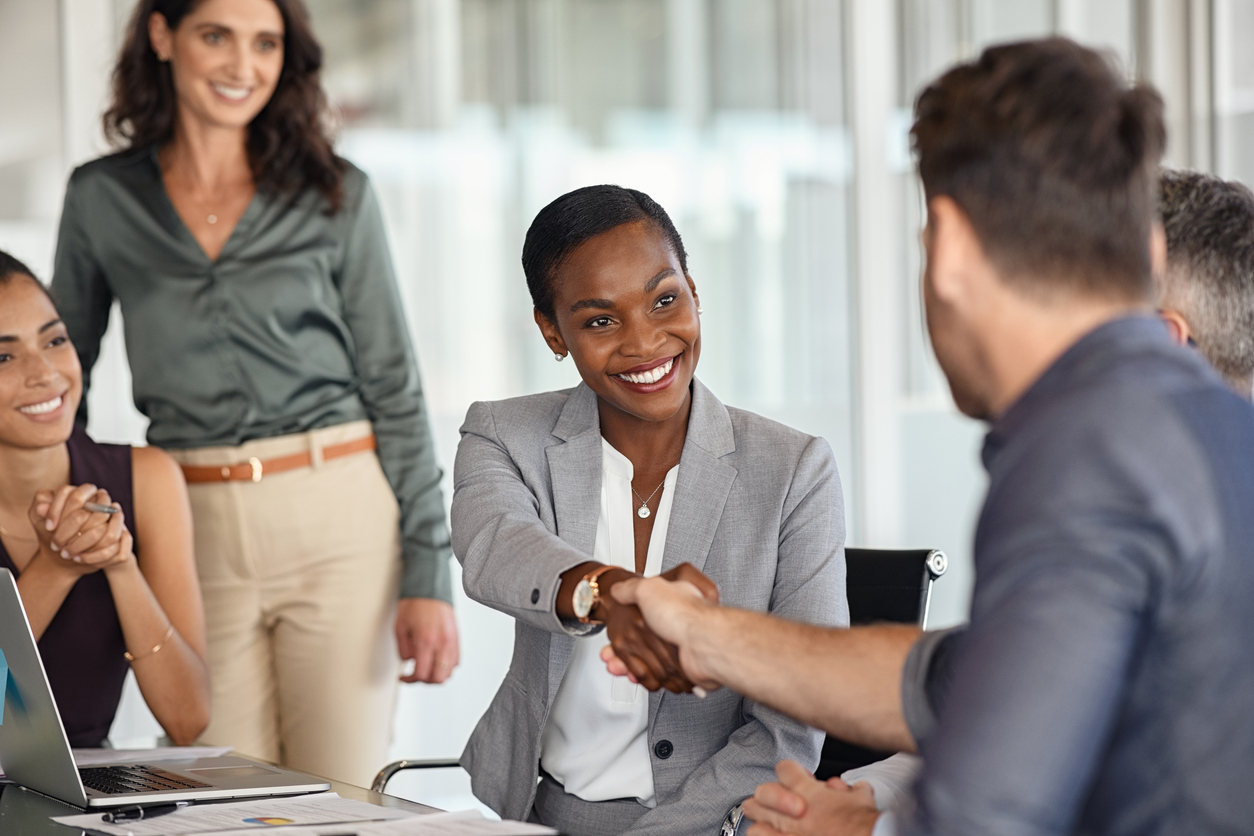 Human Resources Specialist Sitting at a Desk Shaking an Employee’s Hand While Other Employees in the Company Stand Around.